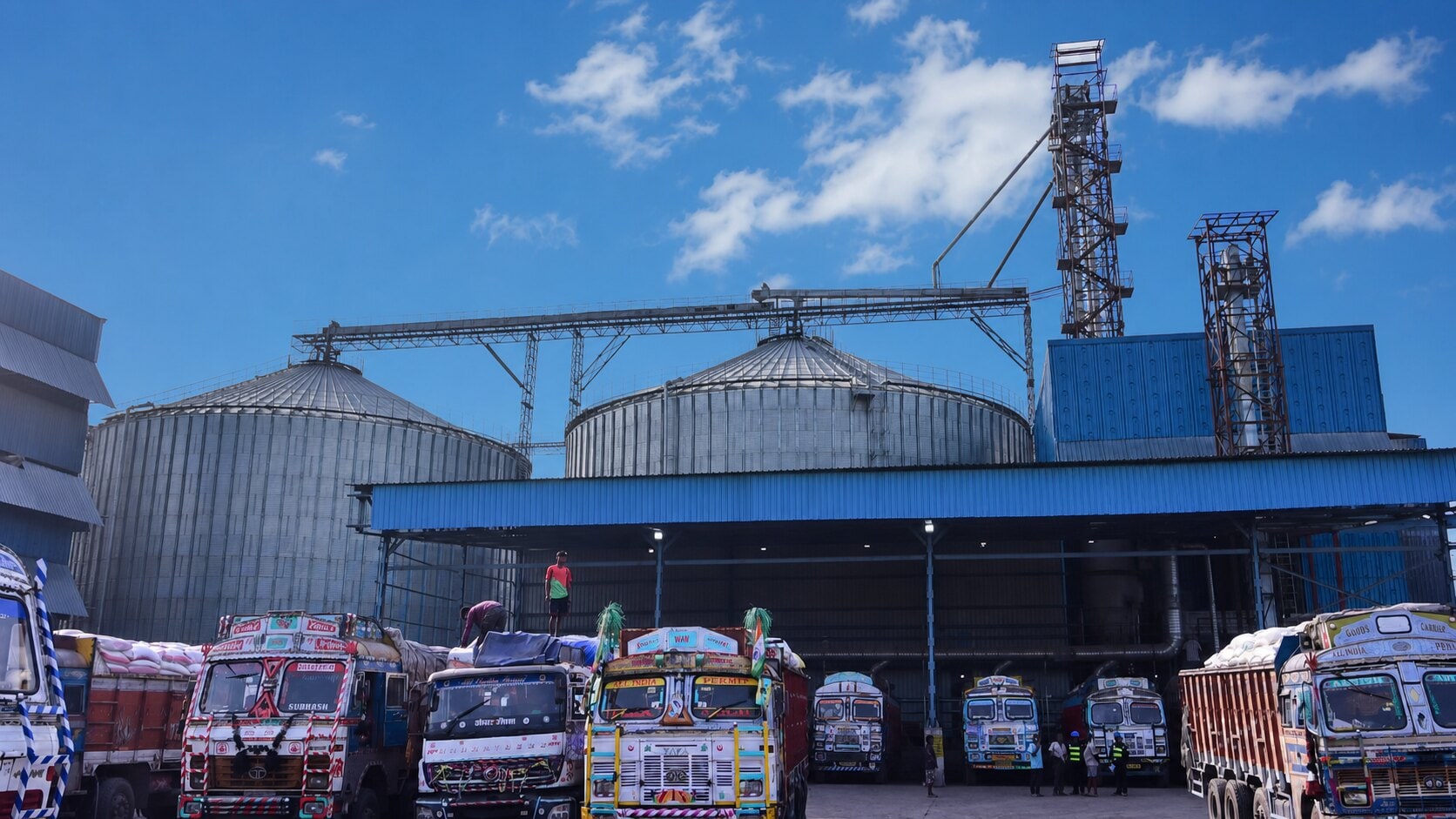 Grain-intake yard at the ethanol plant — corrugated silos, bucket-elevator towers and cargo trucks unloading.
